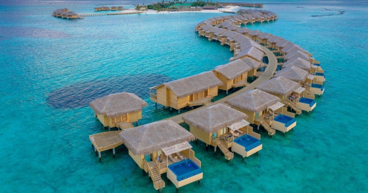 Vista aérea de bangalôs sobre a água cristalina nas Maldivas, conectados por passarelas curvas, cercados por mar azul‑turquesa.