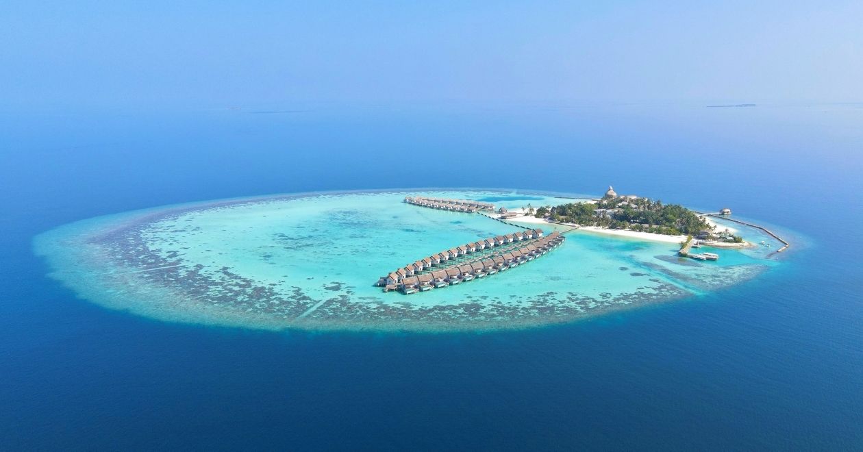 Vista aérea de uma pequena ilha nas Maldivas, cercada por um anel de recife e águas azul‑turquesa, com bangalôs sobre o mar ligados por passarelas e área verde ao centro.
